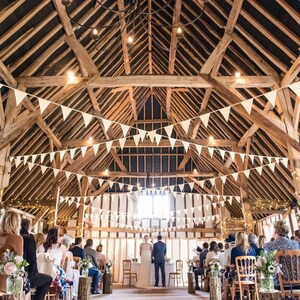 May include: A wedding ceremony inside a rustic wooden barn. White triangular flags hang from the ceiling beams, and string lights are draped throughout. Guests are seated in wooden chairs, facing the couple at the front.