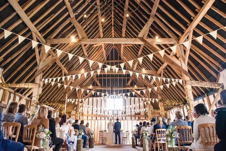 May include: A wedding ceremony inside a rustic wooden barn. White triangular flags hang from the ceiling beams, and string lights are draped throughout. Guests are seated in wooden chairs, facing the couple at the front.