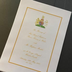 May include: A white wedding invitation with gold trim and a coat of arms featuring a lion and a unicorn. The text reads "The Marriage of His Royal Highness Prince Henry of Wales with Ms Meghan Markle at St. George's Chapel Windsor Castle Saturday, 19th May, 2018 12 Noon."