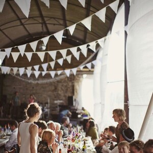 May include: A wedding reception scene with long tables set for guests. White pennant banners hang from the ceiling. Tables are decorated with floral arrangements. People are seated and standing, some are talking and some are eating.