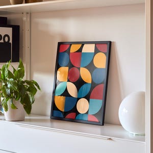 Peut inclure: Une impression géométrique encadrée avec un fond noir et des cercles et des demi-cercles colorés en rouge, jaune, bleu et crème. L'impression est accrochée sur une étagère blanche avec une lampe blanche sur le côté droit.