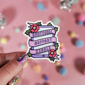 May include: A die-cut sticker featuring a purple e-reader device wrapped in a light pink ribbon with the text "EMOTIONAL SUPPORT KINDLE" printed in black. The design is decorated with green leaves and two pink flowers with dark centres.