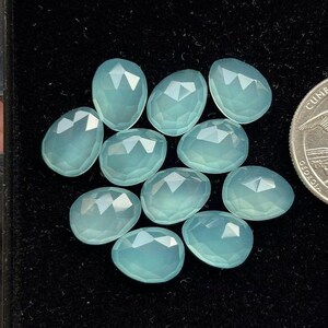 Peut inclure: Dix cabochons de calcédoine bleu clair, chacun avec une forme ovale facetée. Les pierres sont disposées en grappe sur un fond noir.