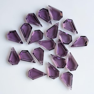 May include: A collection of 20 purple faceted teardrop-shaped glass beads. The beads are scattered on a white surface.