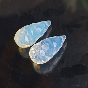 May include: Two teardrop-shaped, iridescent opal beads with a faceted, textured surface. The beads are a pale blue colour with a slight shimmer.
