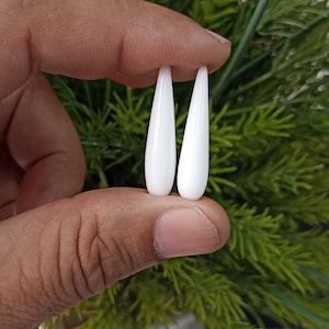 May include: Two white teardrop-shaped beads, held between fingers, against a green leafy background.