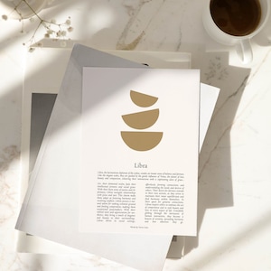 May include: A print with the Libra zodiac sign in gold and text, on a stack of paper. A cup of coffee and flowers are in the background. The print is on a stack of paper.