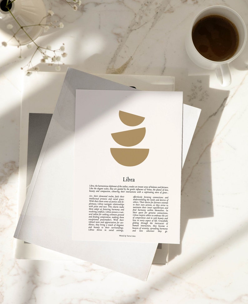May include: A print with the Libra zodiac sign in gold and text, on a stack of paper. A cup of coffee and flowers are in the background. The print is on a stack of paper.