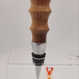 May include: A wooden bottle stopper with a silver metal base and a black rubber seal. The stopper has a unique, textured design and is made of light brown wood.