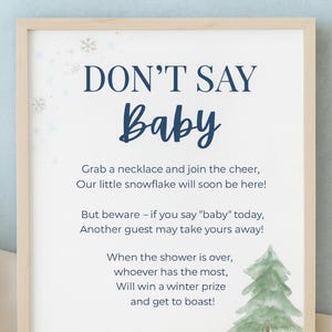 Puede incluir: Un letrero enmarcado para un juego de baby shower con el texto "DON'T SAY Baby". El letrero incluye instrucciones y un tema invernal con copos de nieve y una ilustración de árbol de Navidad. Una cadena dorada en la parte inferior.