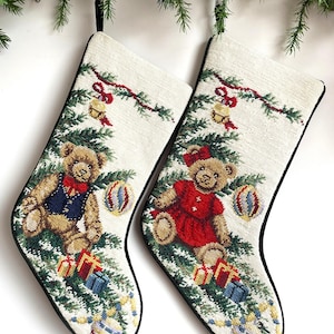 May include: Two cream-colored Christmas stockings featuring a needlepoint design of teddy bears sitting under pine branches. One bear wears a blue vest and red bow tie, the other a red dress with a bow. Both include gifts and ornaments.