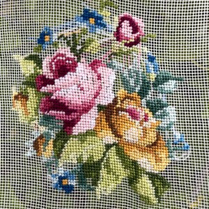 May include: Needlepoint floral design featuring pink, burgundy, and yellow roses, with blue and yellow accent flowers and green leaves. The needlepoint is on a white mesh background, showcasing the detailed cross-stitch work. The design is a classic floral arrangement.