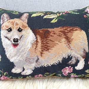 May include: A decorative pillow with a needlepoint design of a brown and white corgi dog on a dark blue background with pink flowers.