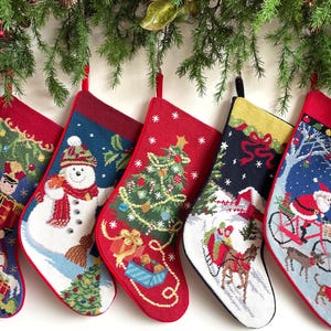 May include: Five needlepoint Christmas stockings featuring festive designs including a nutcracker, a snowman, a decorated Christmas tree, a horse-drawn sleigh in a snowy landscape, and Santa Claus riding a bicycle with a dog nearby.