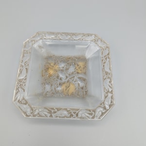 May include: A clear, octagonal glass dish with a decorative border of leaves and vines. The center of the dish features a gold and silver floral design. The dish is likely for decorative purposes.