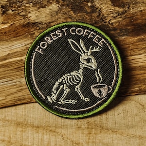 May include: A circular patch with the words "FOREST COFFEE" encircling a skeletal rabbit drinking from a cup. The patch is black with white and green accents, and is likely an iron-on patch or sew-on applique.