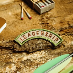 Op de afbeelding: Een tan-kleurig stoffen patch met groene stiksels waarop in bruine letters "LEADERSHIP" staat. De patch is gebogen in de vorm van een boog. De patch ligt op een houten oppervlak met een bruin notitieboek, lucifers en een houten doos.