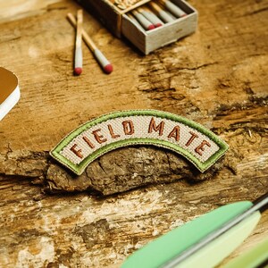 Op de afbeelding: Een bruin leren patch met de tekst "FIELD MATE" in witte letters op een groene achtergrond. De patch ligt op een houten oppervlak met een luciferdoosje en een notitieboek.