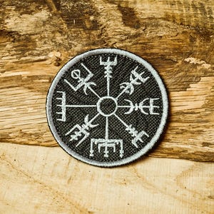 Puede incluir: Un parche bordado en blanco y negro con un diseño de brújula. La brújula tiene ocho puntos y está rodeada por un círculo. El parche está sobre una superficie de madera.