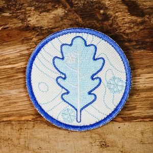 Könnte beinhalten: Ein blauer und weißer gestickter Aufnäher mit einem blauen Eichenblatt-Design auf weißem Hintergrund. Der Aufnäher hat einen blauen Rand und ist von einem weißen Hintergrund umgeben.