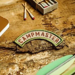 Op de afbeelding: Een groene en bruine geborduurde patch met de tekst "Campmaster" op een houten oppervlak. De patch heeft de vorm van een boog.