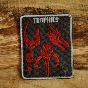 Puede incluir: Un parche bordado negro con la palabra "TROPHIES" en letras blancas. El parche presenta imágenes bordadas rojas de una calavera, un casco mandaloriano y dos calaveras más.