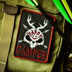 May include: A black and red embroidered patch with the words "HAPPY CAMPER" and a skull design with antlers and arrows. The patch is on a green backpack with arrows.