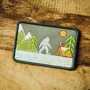 May include: Embroidered patch with a grey background featuring a Bigfoot figure standing in front of a tent and trees. The sun is shining in the sky. The patch has a white border.