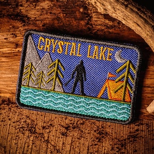 Op de afbeelding: Een blauw en groen geborduurd patch met de tekst "Crystal Lake" en een silhouet van een figuur die voor een tent en bomen staat. De patch heeft een blauwe achtergrond met een golvende groene en blauwe rand.