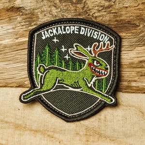 Puede incluir: Parche negro con bordes blancos, que muestra un jackalope verde con dientes y cuernos rojos, sobre un fondo de bosque. El texto "JACKALOPE DIVISION" está arqueado sobre el jackalope.