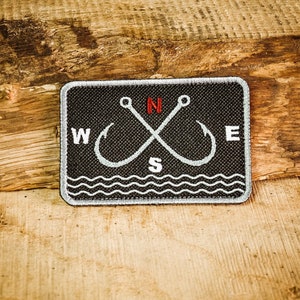 Può includere: Una toppa ricamata in bianco e nero con due ami da pesca incrociati e le lettere N, S, E e W. La toppa è circondata da un motivo a onde bianche.