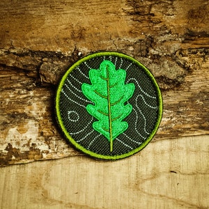 Peut inclure: Un écusson brodé représentant une feuille de chêne verte sur fond noir avec une bordure extérieure verte. L'écusson a un contour blanc et un design texturé.