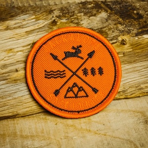 Może przedstawiać: Okrągła pomarańczowa łatka z czarnym haftem. Haft przedstawia jelenia, góry, drzewa, strzały i fale wody. Tekst "Seek Adventure" jest wyszyty wokół krawędzi.