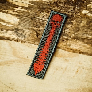 Puede incluir: Un parche negro con un detalle bordado en rojo de una calavera y una columna vertebral.