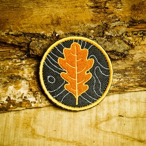 Puede incluir: Un parche redondo bordado en negro y amarillo con un diseño de hoja de roble naranja.
