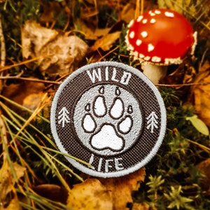 Op de afbeelding: Een zwart-witte geborduurde patch met een pootafdruk in het midden en de woorden "WILD LIFE" rond de rand. De patch ligt op een bed van bruine bladeren en groen mos.