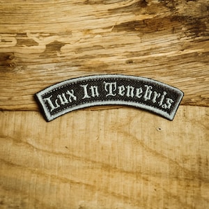 Könnte beinhalten: Ein schwarz-weiß gestickter Aufnäher mit dem Text "Lux In Tenebris" in gotischer Schrift. Der Aufnäher hat eine gebogene Form und einen weißen Rand. Der Hintergrund ist eine Holzoberfläche.