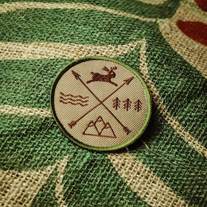 Könnte beinhalten: Ein braunes und grünes gesticktes Patch mit einem Hirsch, Bergen, Bäumen und Pfeilen. Das Patch befindet sich auf einem Jutesack-Hintergrund.