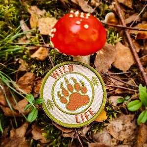 Op de afbeelding: Een ronde, beige patch met groene stiksels en de woorden "WILD LIFE" in groene letters. Een bruine beerpootafdruk bevindt zich in het midden van de patch. De patch ligt op een bed van bruine bladeren en groen mos. Een rode paddenstoel met witte stippen bevindt zich op de achtergrond.