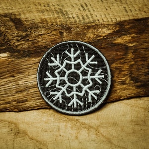 Peut inclure: Un écusson brodé en forme de flocon de neige noir et blanc sur un fond en bois.