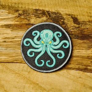 Puede incluir: Un parche circular bordado con fondo negro y un pulpo azul turquesa con ojos amarillos. El pulpo tiene ocho tentáculos y está en el centro del parche.