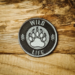 Op de afbeelding: Een geborduurde zwarte en grijze patch met een witte beerpootafdruk in het midden. De tekst "WILD LIFE" is rond de pootafdruk gestikt.
