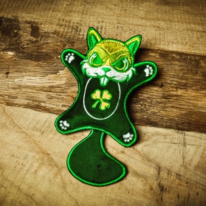 May include: An embroidered patch featuring a cartoon squirrel design in green and yellow, with a shamrock emblem. The patch has white detailing and is set on a wooden surface.