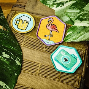 Op de afbeelding: Een bruine canvas tas met drie geborduurde patches. De patches zijn een bierglas, een flamingo en een camera. De tas is een geweldige manier om je persoonlijkheid te laten zien.