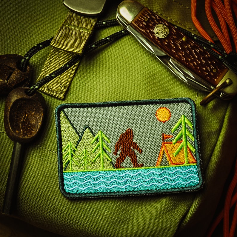 May include: Embroidered patch featuring a Bigfoot design walking near a lake, mountains, and a tent. The patch has a gray background with green, brown, orange, and blue accents. A pocket knife and other camping accessories are in the background.