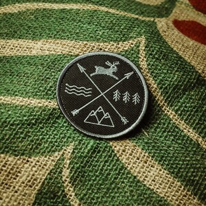 May include: A black embroidered patch with a white outline. The patch features a stylised design of a deer, mountains, trees, and arrows. The patch is on a green and brown burlap background.