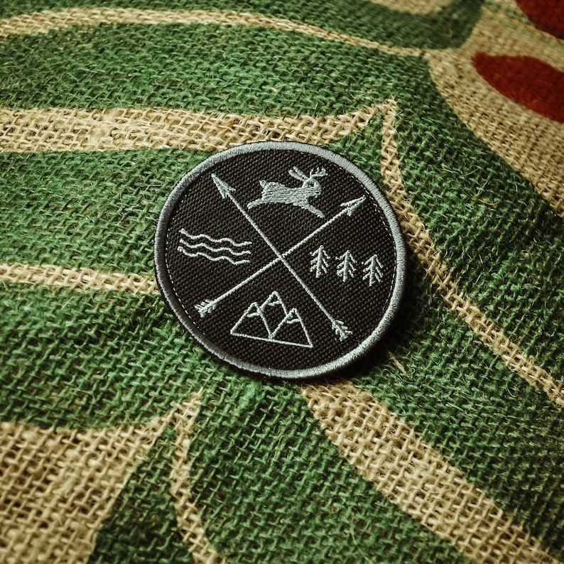 May include: A black embroidered patch with a white outline. The patch features a stylised design of a deer, mountains, trees, and arrows. The patch is on a green and brown burlap background.