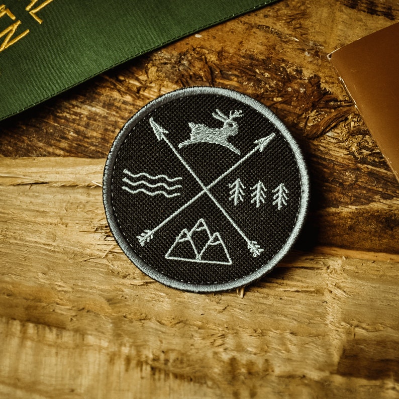 May include: A black and grey embroidered patch featuring a deer, mountains, trees, and arrows. The patch is circular and has a grey border.