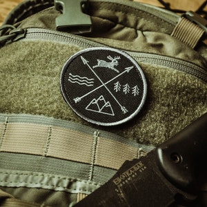 May include: A black and white embroidered patch featuring a deer, mountains, trees, and arrows. The patch is attached to a green and tan rucksack.