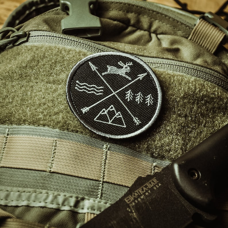 May include: A black and white embroidered patch featuring a deer, mountains, trees, and arrows. The patch is attached to a green and tan rucksack.